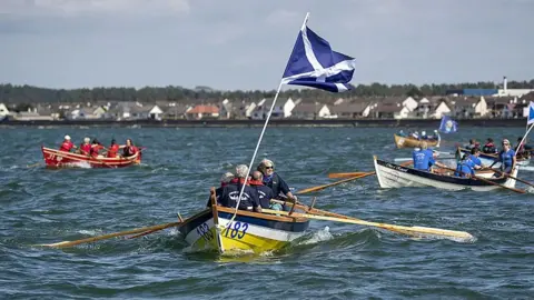Saskia Coulson A blue, white and yellow boat with a Scotland flag on it rows out into the still waters of Loch Ryan