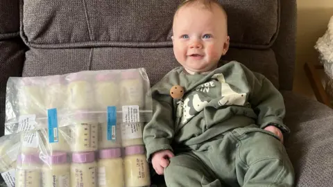 Handout A blond smiling baby boy in a green dinosaur outfit on a grey sofa with a clear bag of thirty or so milk bottles beside him.