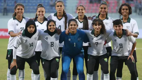 Getty Images The Pakistan women's team posing for a group photo. 10 of the women are in their football kit, which is a white top with black bottoms. The goalkeeper is wearing a light blue kit. Most of them are smiling and you can see them on the football pitch with the stadium in the background. 