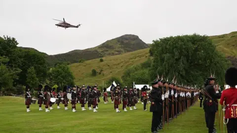 PA Media Lines of soldiers and military personnel wearing uniforms in the gardens on the Palace of Holyroodhouse. Arthur Seat is in the background and a helicopter is in the sky. 