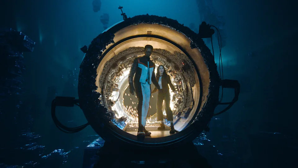 A Black man in a white, black and red superhero costume is stood inside a large circular machine. that is glowing. On his right there is a woman with dark hair wearing a dark jacket, white t-shirt, and dark trousers. There is a black material growing inside the machine. Outside is a dark mysterious atmosphere.