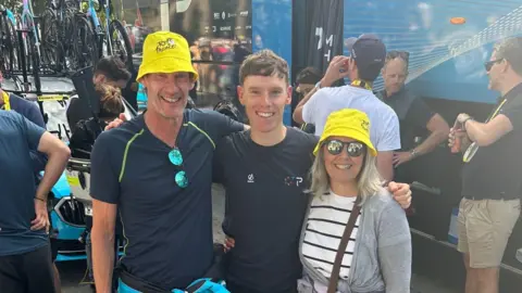 BBC Two men in dark blue tshirts and a woman in a white and black striped shirt and grey cardigan are standing in a line with their arms around each other and are smiling at the camera. The man and the woman on each end of the row are wearing yellow bucket hats