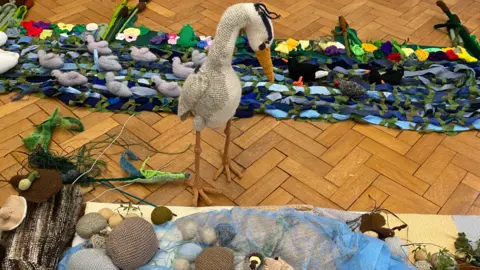 The Secret Society of Hertford Crafters A heron, looking at a river scene, all made out of fabric, the heron is standing up.
