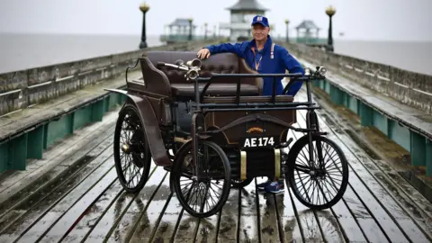 Mark Reber A rainy Clevedon Pier, with Mark Reber standing beside a brown wooden framed Victorian motor car