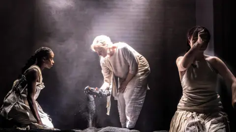 Raisa Šehu A scene in a play with three actors dressed in minimalist, loose clothing. A woman in the left is crouched watching another woman pulling a sandal from a piles of black dust. To her right another woman is holding an outstretched fist to the camera.