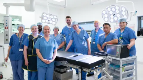 Leeds Teaching Hospitals Trust 10 medics, all of them wearing blue gowns pose next to a bed in an operating theatre.