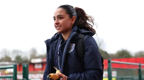 Getty Images Mariam is smiling with a banana in her hand. She has a navy winter jacket on. In the background you can see a football pitch.