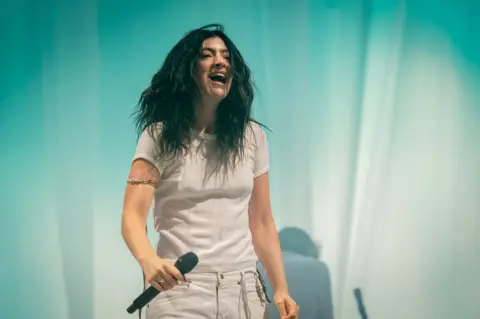 Getty Images Singer Lorde wears a white t-shirt and trousers in front of a turquoise curtain on-stage. She's holding a microphone and smiling.