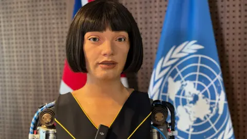 Copyright 2025 © Ai-Da Robot Studios The ultra-realistic humanoid robot artist Ai-Da speaking to the camera. The United Nations flag is behind her.
