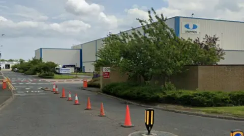 Google A grey and blue factory building sits on the right hand side of a road. A wall covered by trees and bushes can be seen in front of it, to the right. There are orange traffic cones laid out on the road.