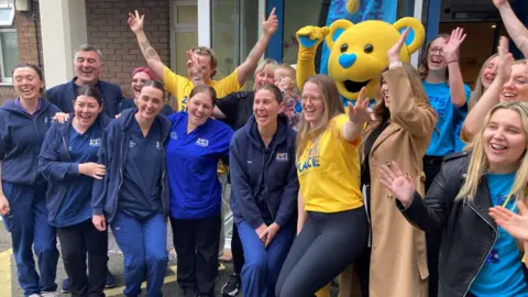 Tom Walker/BBC A group of nurses, some in uniform, and fundraising staff in blue-and-yellow Zoe's Place branded t-shirts, as well as a mascot in a yellow teddy-bear costume, smile and cheer. Liverpool West Derby MP Ian Byrne is standing at the back left of the group as we see it.
