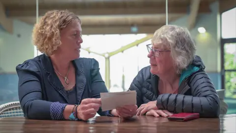 BBC News Two women sat at a table next to each other, looking at family photographs. They look at each other as they hold the photos.
The woman on the left is wearing a navy jacket and has short ginger curly hair. The women on the right has short grey hair, glasses and is wearing a dark coat.