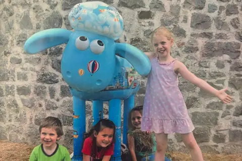 Naomi/Family copyright A younger Naomi, aged five, wears a pink dress and stands beside a pale blue Shaun the Sheep sculpture decorated with colourful hot air balloons on its face. Three other children — her siblings and a friend — are sitting under the sculpture, smiling for the camera.
