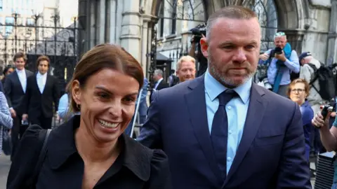 Reuters Coleen Rooney smiling next to Wayne Rooney, both walking away from court with photographers in the background on 17 May 2022