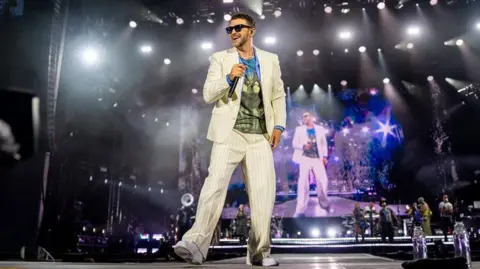 CUFFE & TAYLOR Justin Timberlake is standing in the middle of the picture on a stage. He is looking to the left of the camera and is wearing a matching cream striped suit, with a blue and green T-shirt. He is holding a microphone and there are backing singers behind him. 