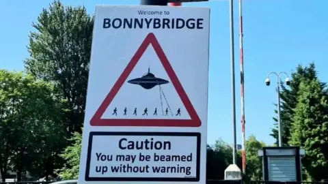 @ASignOfHumour A sign in Bonnybridge which shows a UFO zapping tiny humas. The sign says Caution: You may be beamed up without warning