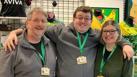 Chris Goreham/BBC Members from the On The Stall City shop standing in a row looking directly at the camera and smiling. Andy Bowles is on the left dressed in a black T-shirt and grey zip up sweatshirt. He is wearing a pair of glasses and has a green lanyard round his neck. 