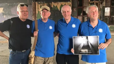 Four men stood in a line. They are all wearing tops with a circle blue circle club logo on the chest. Dave, on the far right, is holding an old black and white photo of the Blue Moon Club from the 1960s
