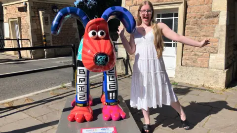 BBC Naomi, aged 17, stands smiling beside a brightly coloured Gromit sculpture. She is wearing a white dress and striking the same playful pose she did as a young child. The sculpture features vivid patterns and cheerful colours.