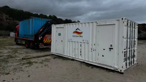 Carlyon Bay Surf Life Saving Club A white shipping container with a sign attached to it. There is a lorry with a blue container on the back.