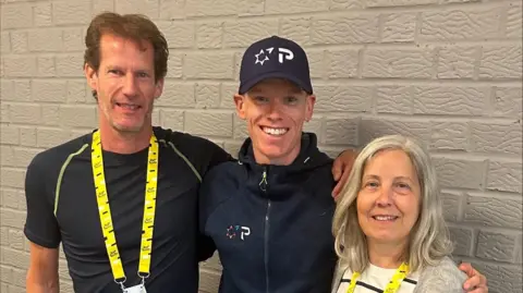 Two men in dark blue tshirts and a woman in a white and black striped shirt and grey cardigan are standing in a line with their arms around each other and are smiling at the camera. The man and woman on either end of the row are both wearing yellow lanyards while the man in the middle is wearing a cap