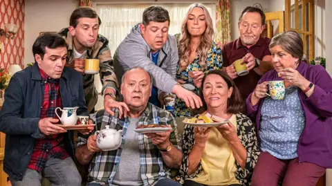 BBC Eight people gathered around a sofa in a living room. Some of them are carrying a selection of mugs, sandwiches and cakes. Two people are on the sofa, two on the arms of the sofa and four are standing behind with two of them reaching out to the plates of food