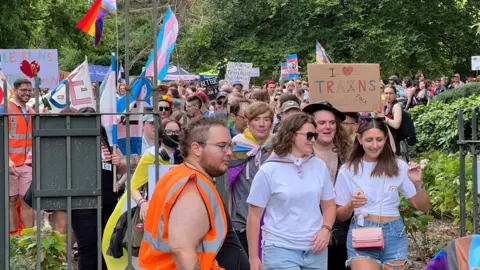 BBC A large crowd of people walking through the metal gates of an outdoor park during the Pride event in Bath. Many people are holding up pride flags or handmade signs expressing support for LGBTQ+ rights.