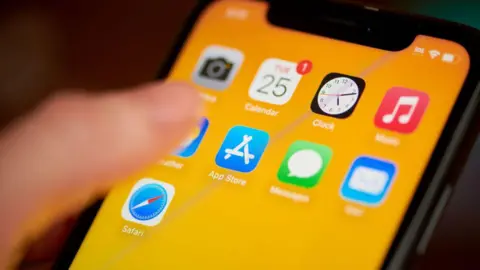 Getty Images Someone's thumb hovers over the App Store widget on an iPhone screen, which has a bright orange wallpaper. The App Store app is surrounded by other Apple app icons on the screen.