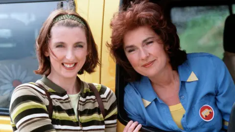 BBC Julie Wilson Nimmo smiles alongside Juliet Cadzow who leans out the window of a yellow bus