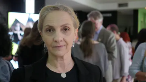A lady with white blonde hair looks into the camera, half smiling. She is wearing a black dress and blazer, as well as silver jewellery.