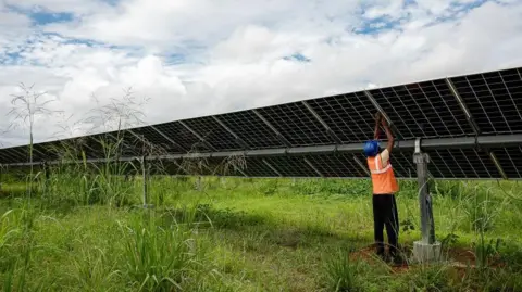 Getty Images A man in hi vis and a hard hat reaches up to a large solar panel in a field.
