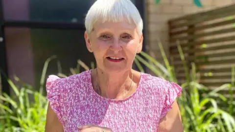 North West Anglia NHS Foundation Trust Rosie Collcott has short cropped fair hair with a fringe and is wearing a pink and white print dress. She is standing in the sun with plants and a fence behind her.