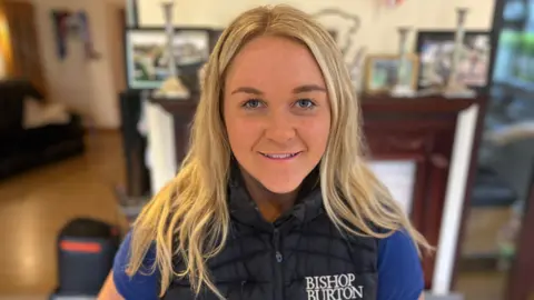 BBC A young woman with long, blonde hair smiles at the camera. She is wearing a  dark blue padded gilet with a blue T-Shirt underneath