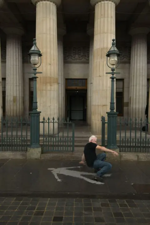 Andy Goldsworthy Andy Goldsworthy is got in mid action from getting up from the wet ground. There is an imprint of his body shame on the pavement that is dry.