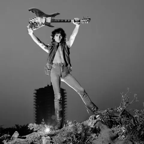 Getty Images A black and white image of Robin George on top of a rocky outcrop holding his guitar aloft. He is wearing a short-sleeved jacket, jeans and boots and is bare chested.