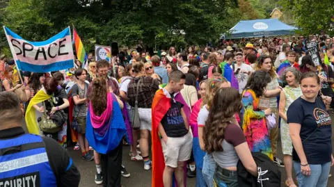 A large crowd of people gathering in an outdoor park for the Pride event in Bath. Many of them are draped in pride flags, and several are holding up signs in support of LGBTQ+ rights.