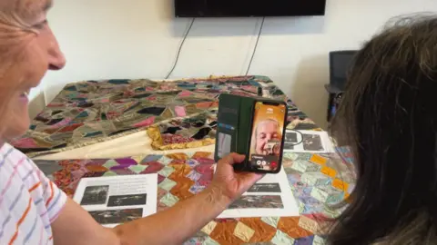 Aisha Iqbal/BBC Two women on a video call with a third woman. In front of the group is a table with a patchwork quilt laid on top of it, as well as various bits of paper with words and pictures on them.