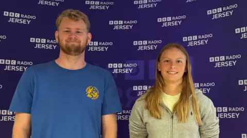 Josh Twohig-Jones on the left wearing a blue top with a Jersey Youth Service badge, and Lauren wearing a grey hooded jacket. They are both smiling at the camera and stood in front of a purple Radio Jersey sign.