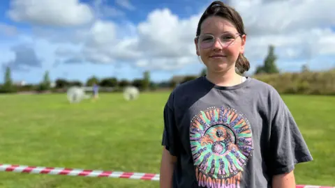 Orla stands on a grassy field under a blue sky with scattered clouds. Their face is blurred for privacy. They are wearing a dark gray t-shirt with a colorful psychedelic design of the sun and moon, and the word 'MOONLIGHT' at the bottom. In the background, two large inflatable balls and red-and-white striped tape are visible.