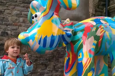 Barney/Family copyright Three-year-old Barney reaches up toward a Gromit sculpture. He wears a red T-shirt under a bright blue raincoat with multicoloured details. Gromit is painted in a mix of vivid colours and patterns.