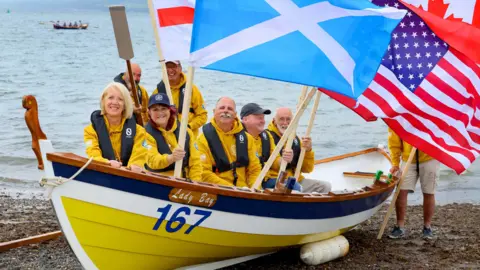 Colin Hattersley A boat crew dressed in bright yellow wave a range of flags while sitting in a yellow, white and blue boat on the south of Scotland shoreline