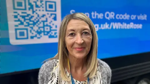 BBC News A woman with long blond hair and wearing a grey jumpers smiles as she stands in front of a blue van. On the side of the van is displayed a large white QR code  and text (partially obscured) reading, "Scan the QR code or visit ycr.org.uk/whiterose".