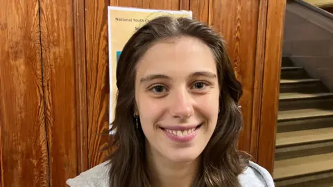 BBC Amy Patton has long brown wavy hair and brown eyes. She is smiling at the camera in front of a brown wooden wall and cream staircase.