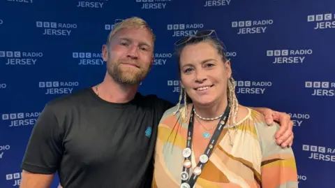 BBC Will Crawfrod and Rachel Boss stand side-by-side, smiling for the camera in a radio studio. Will has blonde hair and a beard, and is wearing a dark t-shirt. Rachel, has dark hair with some braids and is wearing a colourful patterned top, with a lanyard around her neck displaying several buttons. Behind them is a dark blue wall with "BBC RADIO JERSEY" printed repeatedly in white.