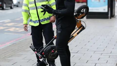 pa Anonymous shot of a person wearing a black tracksuit and black gloves carrying a folded e-scooter talking to a police officer who is wearing black trousers and a reflective jacket. They are stood on the pavement and the shot shows the middle of their bodies only.