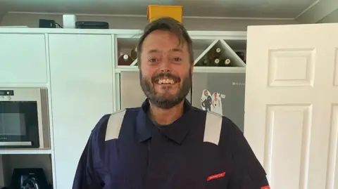 Anna-Louise Stubbings Zach standing in a kitchen wearing a blue military boiler suit and smiling at the camera. He has a short beard and brown hair.
