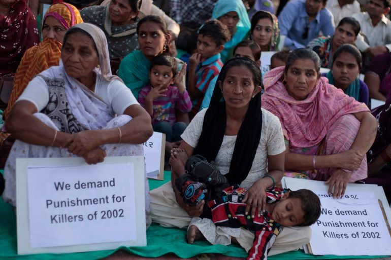Protesters in Gujarat sit on the ground with protest signs that read "We demand punishment for the killers of 2002"