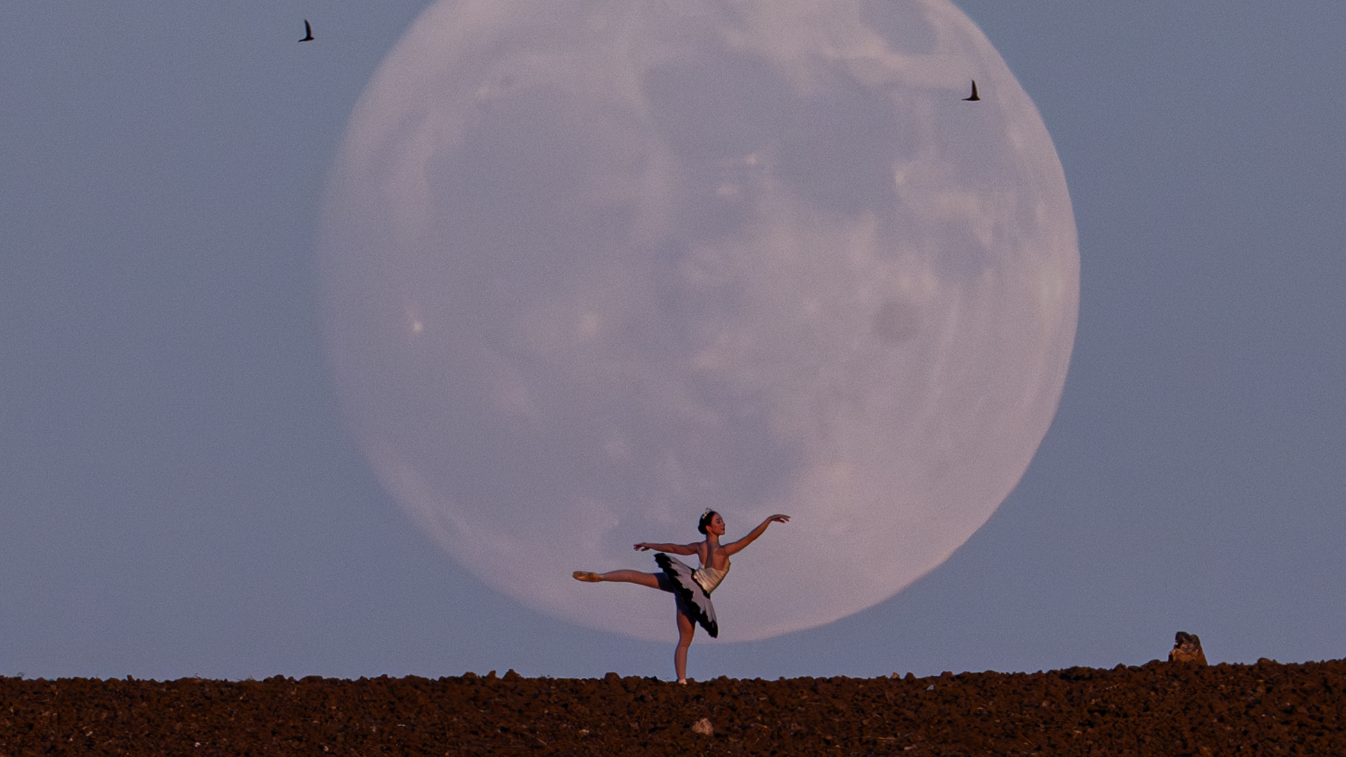 A ballerina is seen striking a pose with a large full moon looming behind her in the evening sky.
