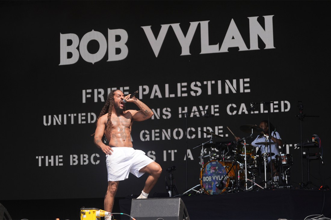 Bob Vylan wearing white shorts and no shirt singing into a microphone while lifting one leg in preparation to stomp on stage at Glastonbury.