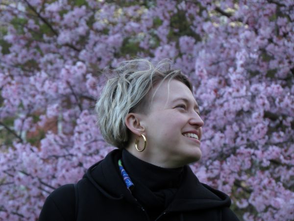 A smiling young woman with short blonde hair, in front of a hedge covered with purple flowers.